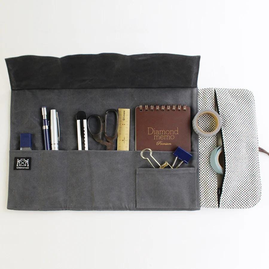 A gray fabric pouch with various office supplies and a notebook inside, including pens, scissors, and a stapler.