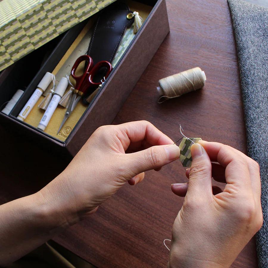 A person's hands are carefully threading a needle on a wooden surface, surrounded by various crafting supplies such as scissors and a spool of thread.