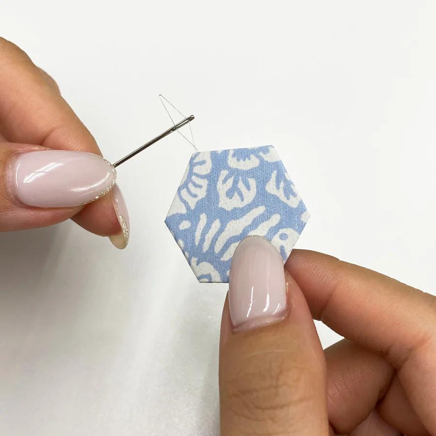 A pair of hands holding a needle and thread, with the needle piercing through a blue and white hexagonal patterned fabric.
