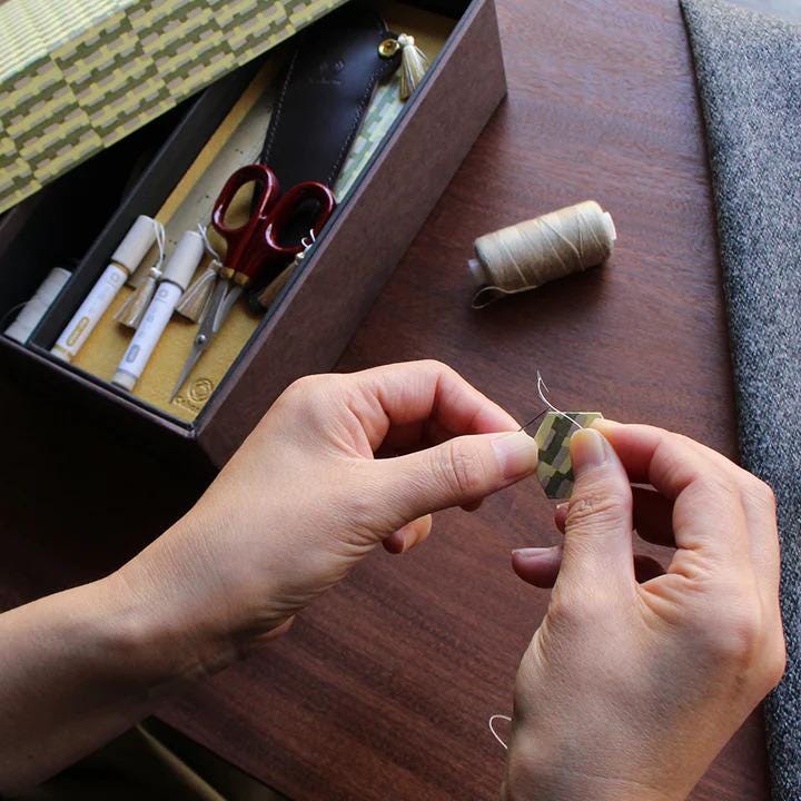 A person's hands are carefully threading a needle with a piece of thread on a wooden surface, surrounded by various crafting supplies.