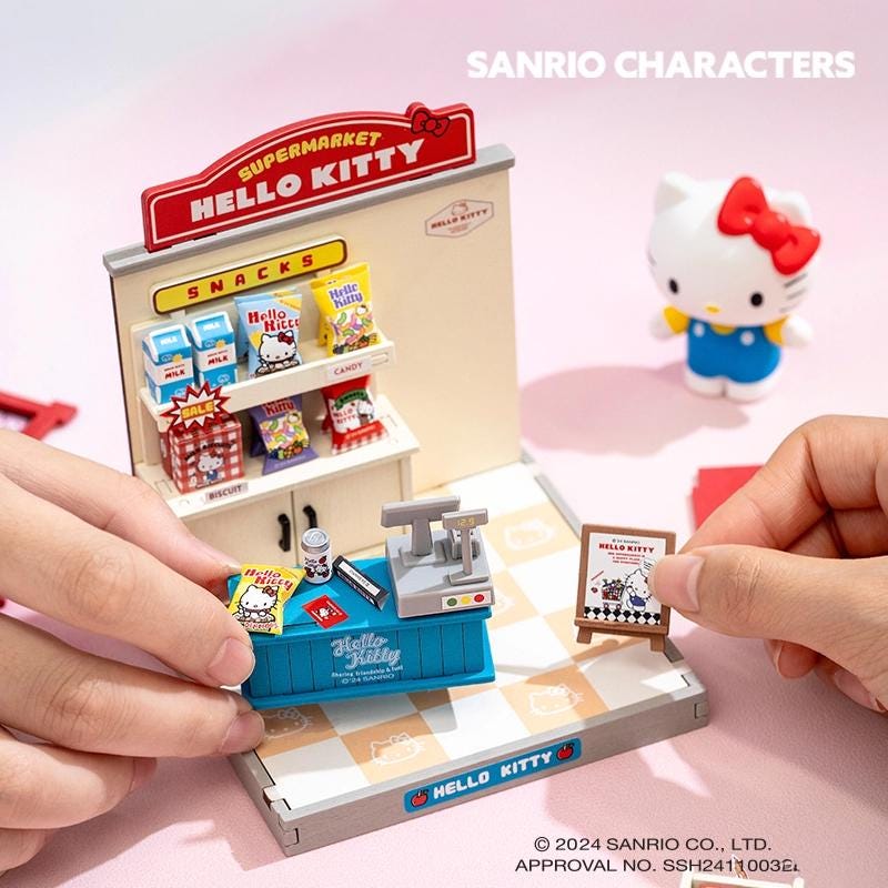 A person is holding a Hello Kitty miniature grocery store set, which includes a Hello Kitty cash register and various Hello Kitty snacks and candies.