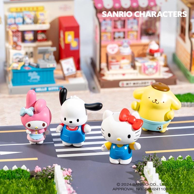A group of four Hello Kitty figurines, including a pink one, a black and white one, a yellow one, and a red one, standing on a miniature street scene with a toy store in the background.