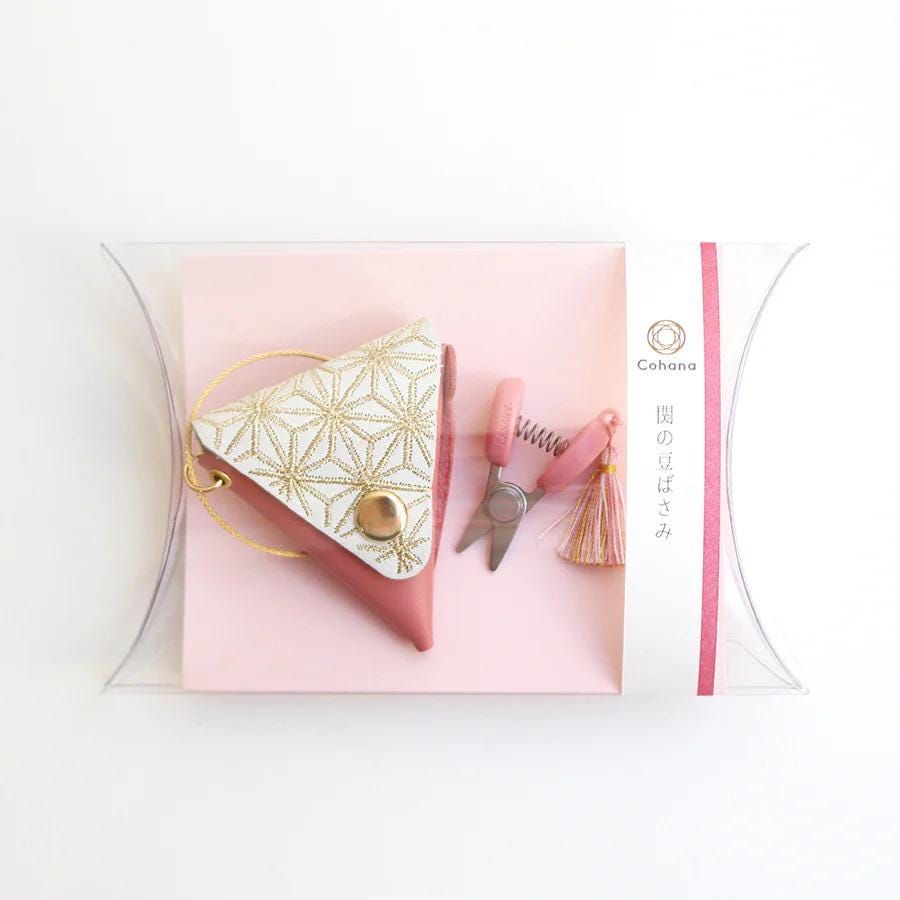 A pink and white geometric-patterned purse with a gold button, a pair of pink scissors, and a pink tassel are displayed on a pink background.