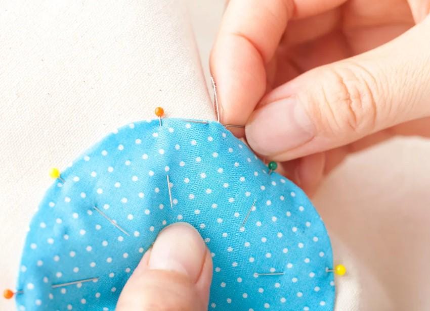 A hand is carefully sewing a blue fabric with white polka dots, using a needle and thread.
