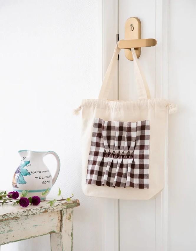 a white door with a gold door handle, a white tote bag with a checkered pattern hanging on the door, and a white pitcher with a blue design on a white table.