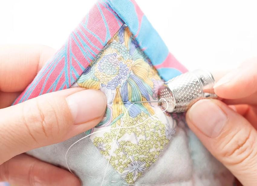 A pair of hands carefully sewing a colorful quilt with a thimble and needle.