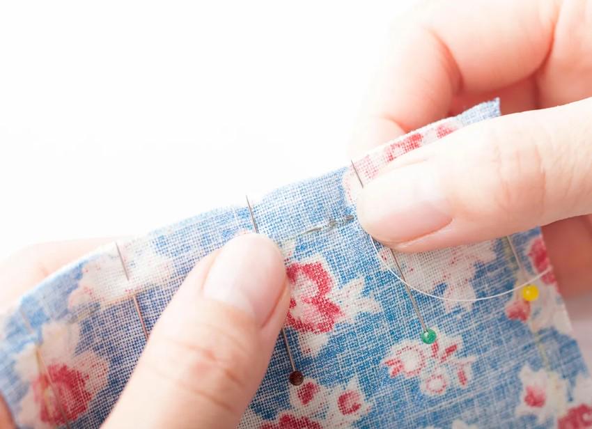 A person's hands are shown sewing a piece of fabric with a floral pattern, using a needle and thread.