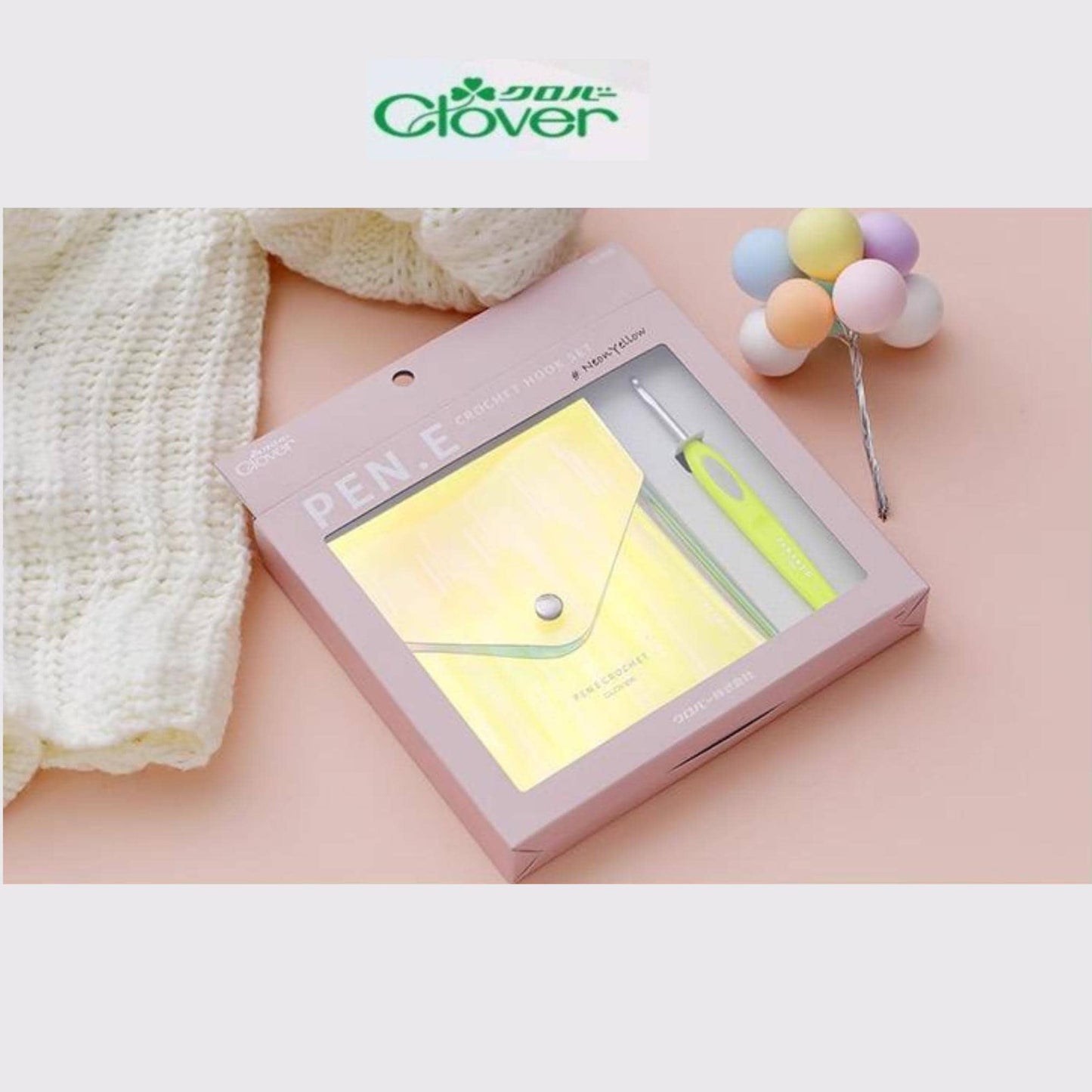 A pink box containing a pen and a crochet hook, along with a crocheted ball and a white knitted item, all placed on a pink surface.