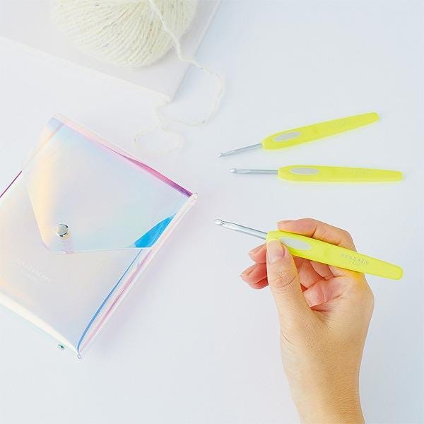 A hand holding a yellow crochet hook, with a small white purse and a ball of white yarn visible in the background.