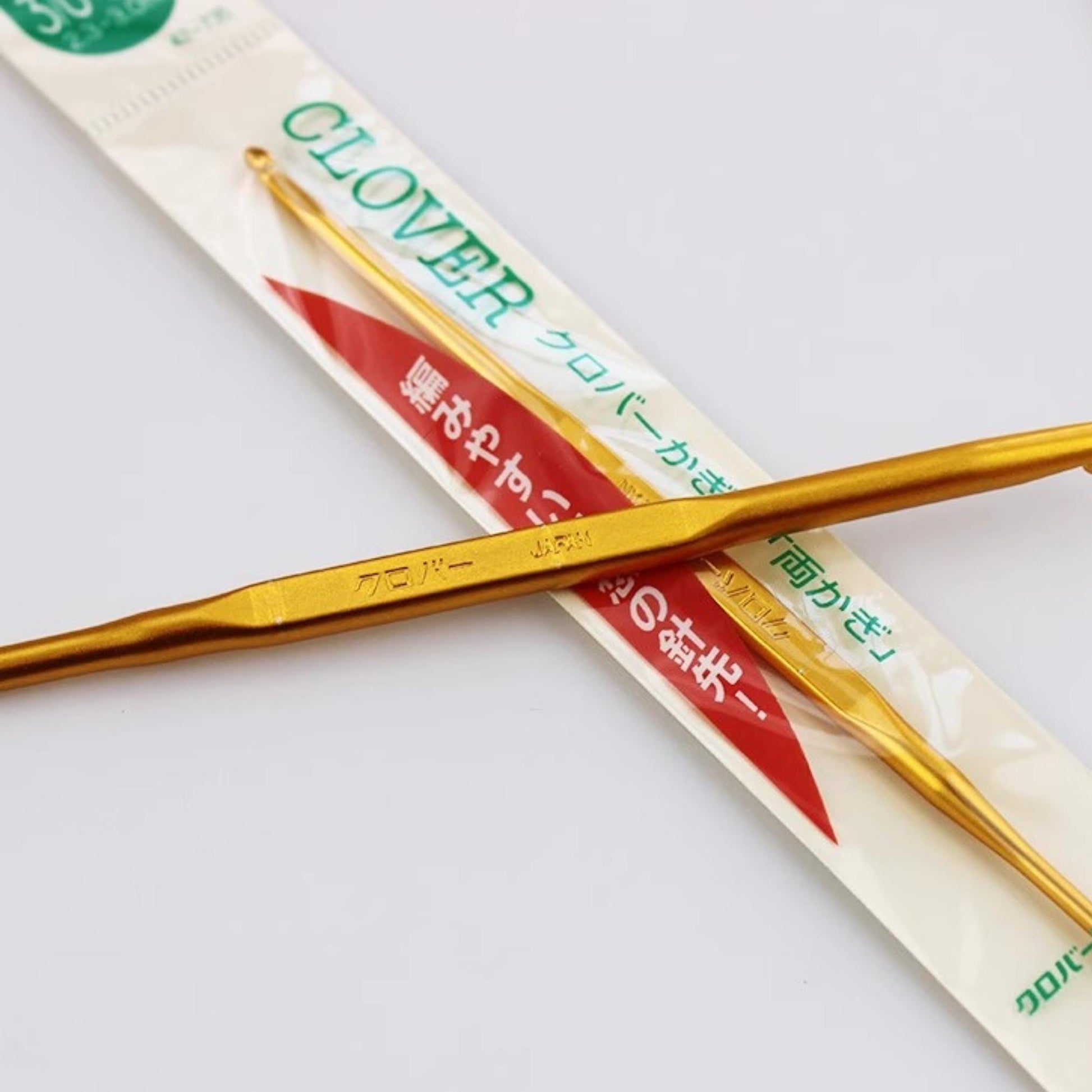 a pair of gold crochet hooks crossed over each other, with a package of the same type of crochet hooks visible in the background.