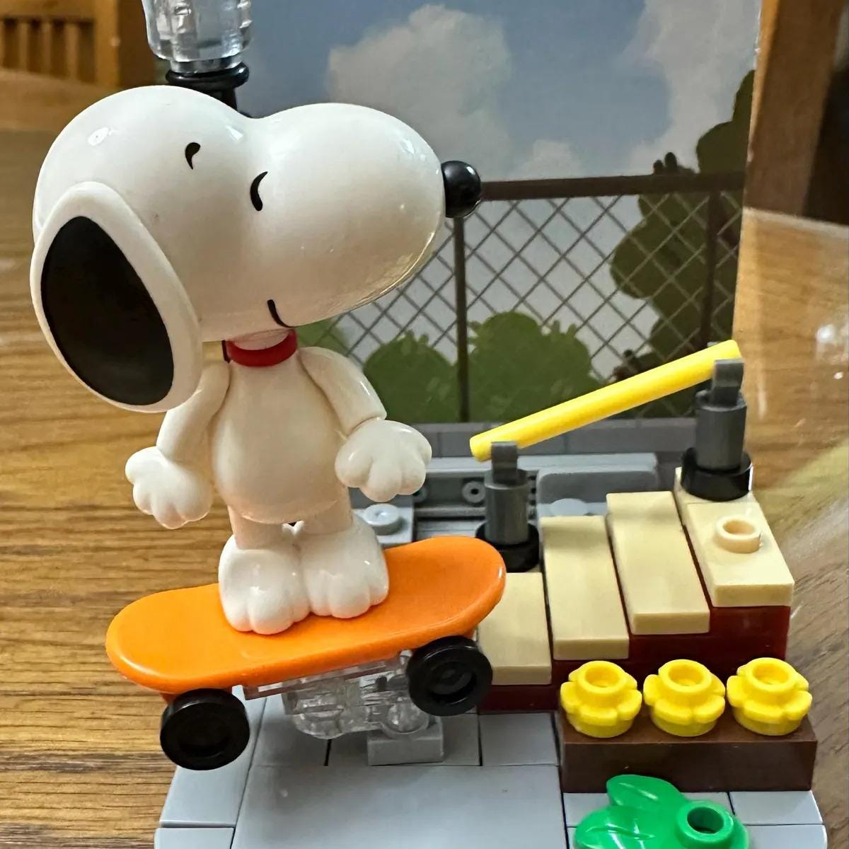 A white toy dog, resembling Snoopy from the Peanuts comic strip, is standing on an orange skateboard and appears to be riding it. The scene includes a toy fence and other small toy objects, creating a playful and imaginative setting.