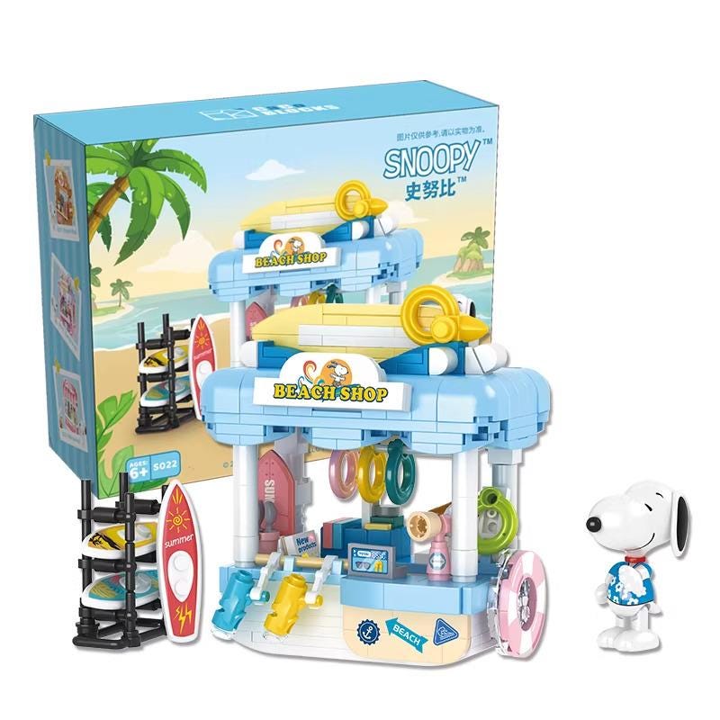 A toy set depicting a beach shop with a Snoopy character standing next to it. The toy set includes a blue and yellow building, surfboards, and other beach-themed accessories.