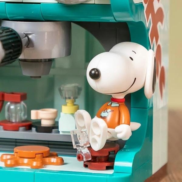 A toy figure of a white dog wearing an orange shirt is sitting on a toy coffee machine.