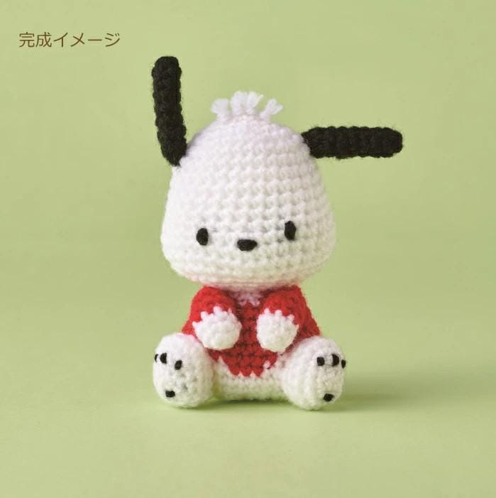 A crocheted toy resembling a small dog or puppy, with a white body, black ears, and a red shirt.