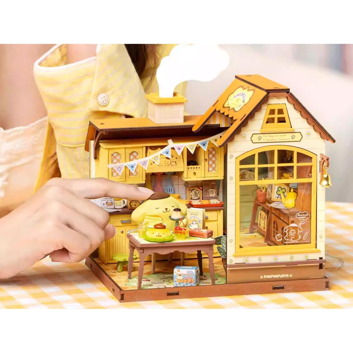 A person's hand is holding a small wooden dollhouse with a yellow and brown color scheme, featuring a kitchen area with a table and chairs.