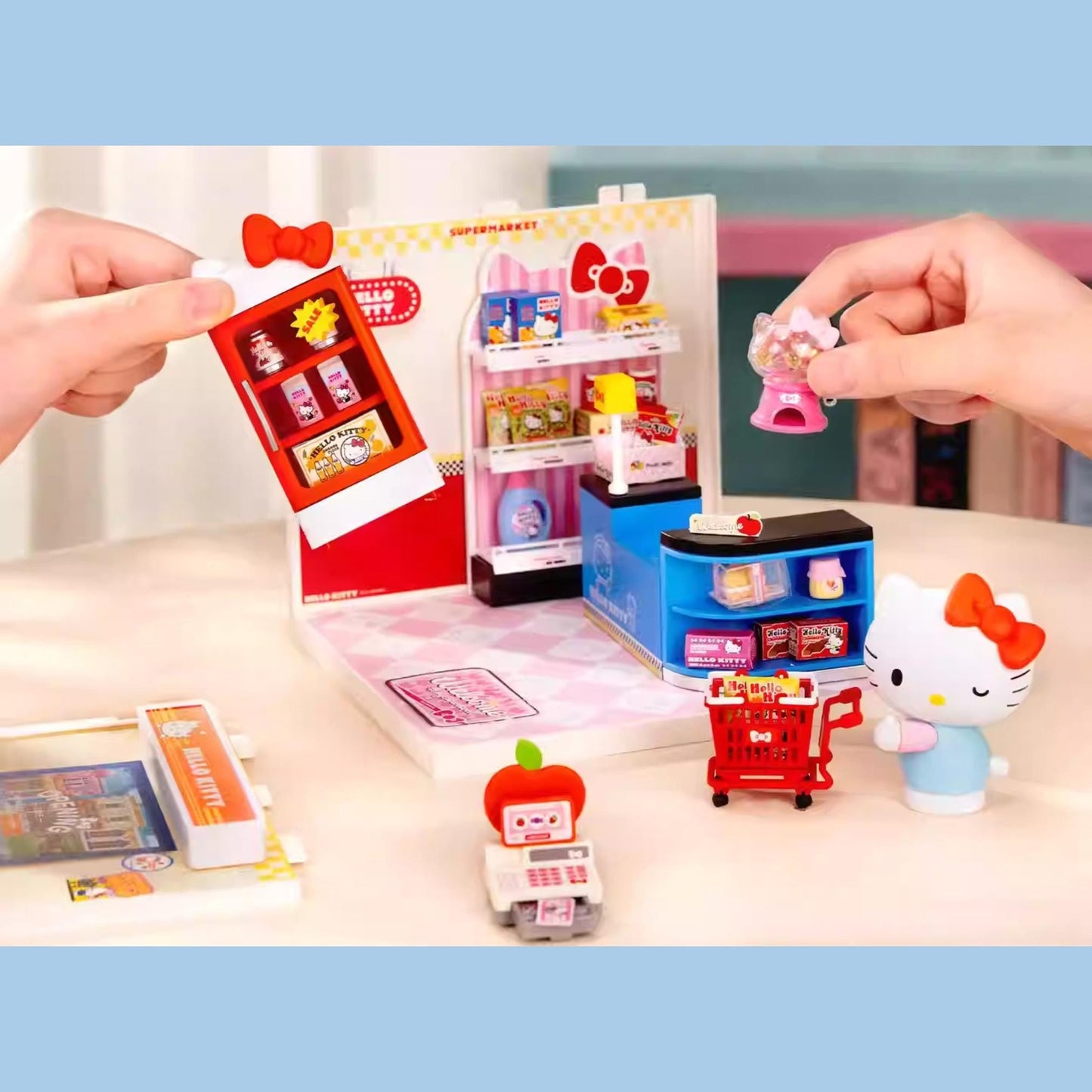 A person's hands are holding up a Hello Kitty toy store set, which includes a toy cash register, a toy apple, and other Hello Kitty-themed items.