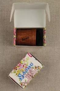 a wooden object, possibly a pen or pencil holder, placed inside a white box with a floral pattern. The box is open, revealing the wooden object inside.