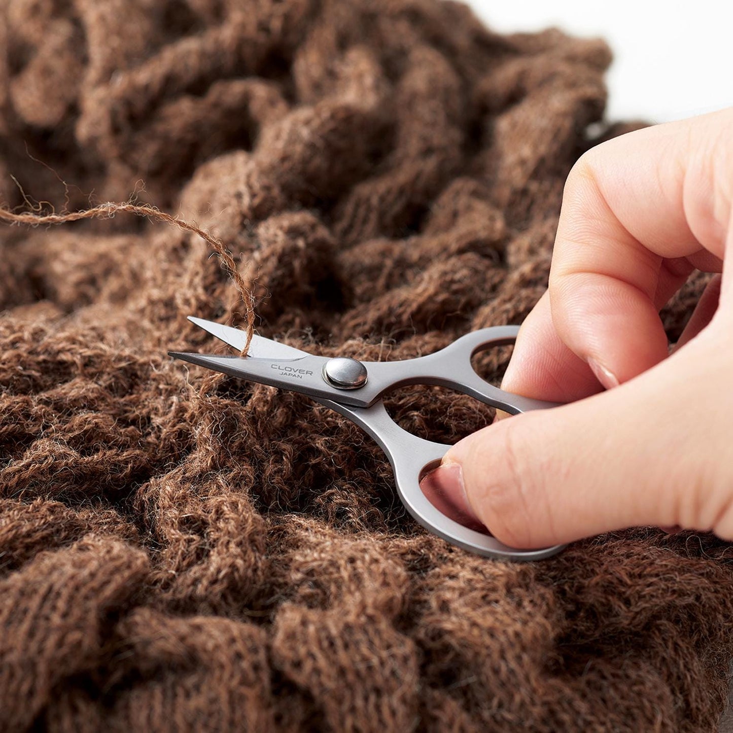 A hand is holding a pair of scissors and cutting a piece of brown yarn.