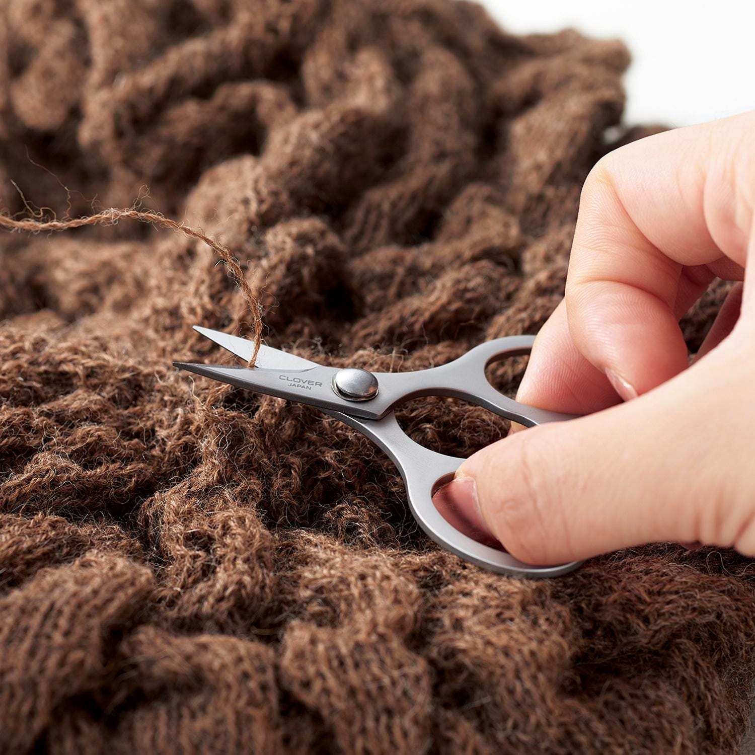 A hand is holding a pair of scissors and cutting a piece of brown yarn.