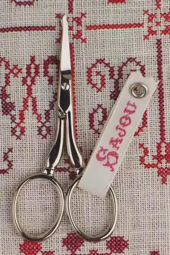 A pair of silver scissors with a white tag attached to them, resting on a red and white embroidered fabric.