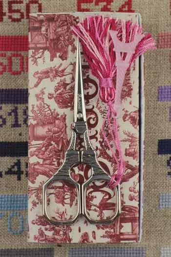 A pair of silver scissors with a pink tassel is displayed on a patterned box.