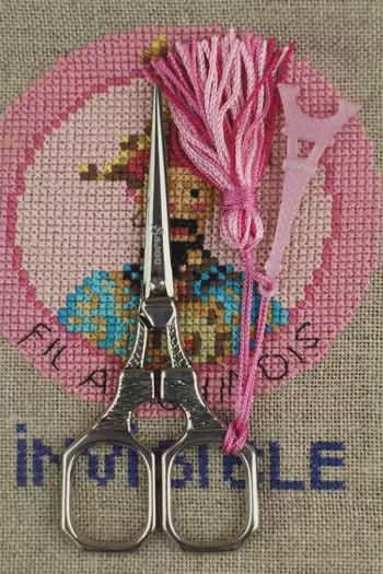 a pair of scissors with a pink tassel attached to them, placed on a pink and beige fabric with the word "INVICTA" written on it.