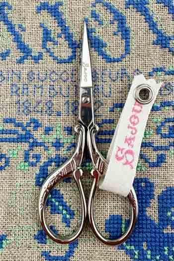 A pair of silver scissors with a white tag attached to them, resting on a blue and green patterned fabric.