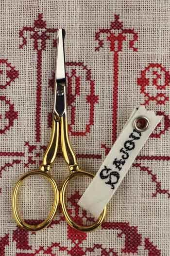 A pair of gold-colored scissors with a white tag attached to them, placed on a red and white embroidered fabric.