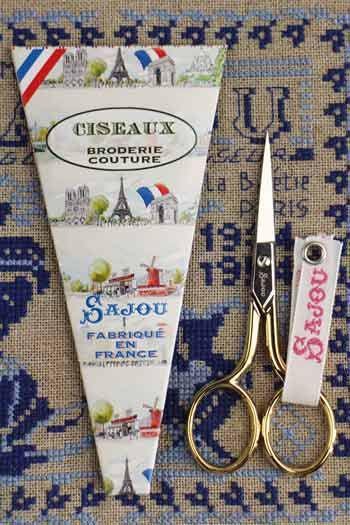 a pair of gold-colored scissors and a white paper with a French-themed design, including illustrations of landmarks and text.