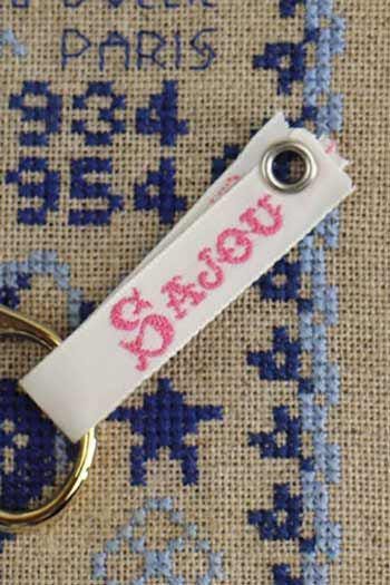 a keychain with a white strap and a metal ring, attached to a beige fabric with a blue and white embroidered pattern.