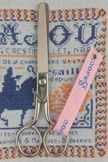 A pair of scissors and a pink tag with the word "DOU" printed on it are placed on a cross-stitch pattern.