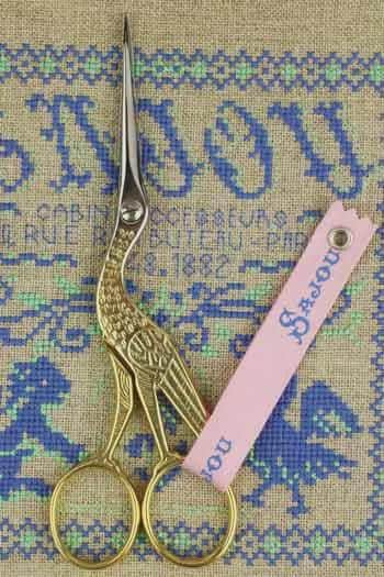 A pair of gold-colored scissors with a pink tag attached to them, placed on a beige fabric with blue and green embroidered designs.