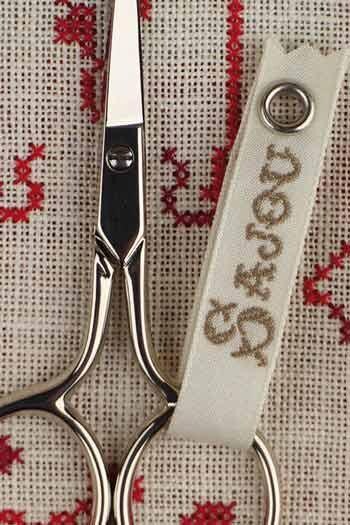 A pair of silver scissors with a white tag attached to the handle, resting on a beige fabric with red embroidered patterns.