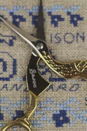 A close-up image of a pair of gold-colored scissors with a cross-stitch pattern in the background.