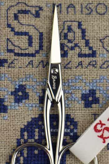 A pair of silver scissors with a label on the handle, placed on a cross-stitch pattern with blue and white designs.