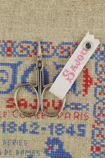 A pair of scissors with a white tag attached to them, resting on a beige fabric with a cross-stitch pattern.