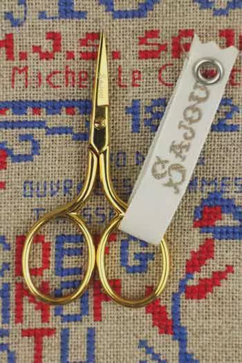 A pair of gold-colored scissors with a white tag attached to them, resting on a beige fabric with a blue and red embroidered pattern.