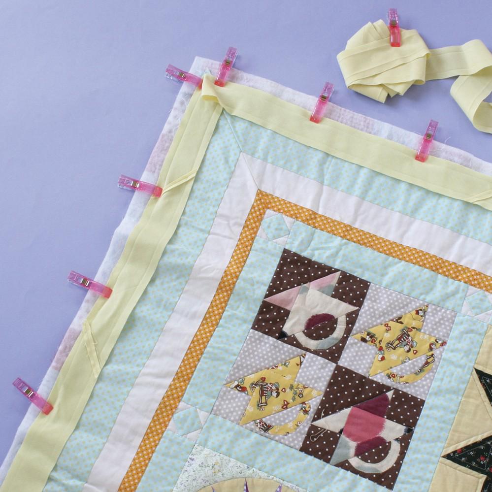 a colorful quilt with a yellow border, featuring a variety of patterns and designs. The quilt is being secured with yellow fabric and plastic clips, indicating that it is being prepared for sewing or assembly.