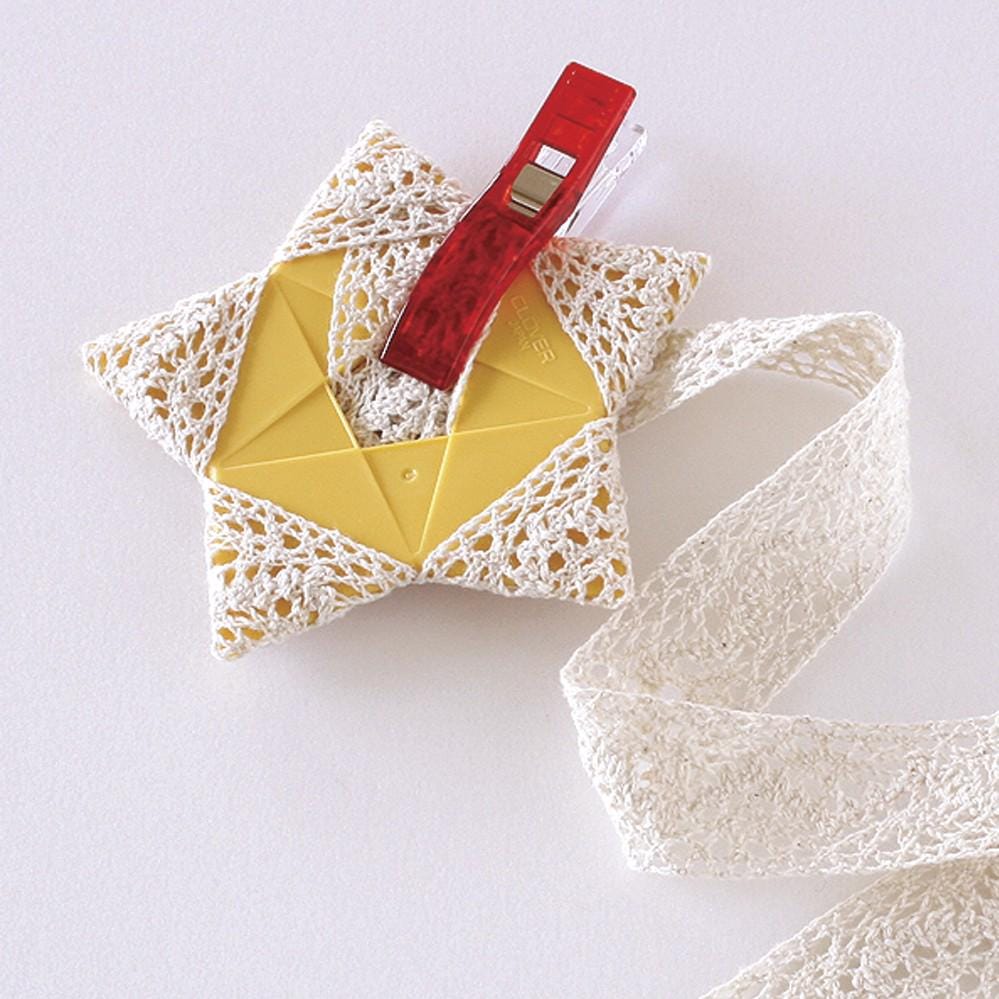 A crocheted star-shaped object with a red plastic clip attached to it, placed on a white surface next to a white lace ribbon.