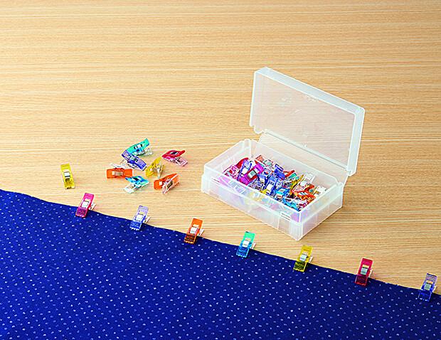 A clear plastic container filled with colorful sewing clips is placed on a wooden surface, with a blue fabric nearby.