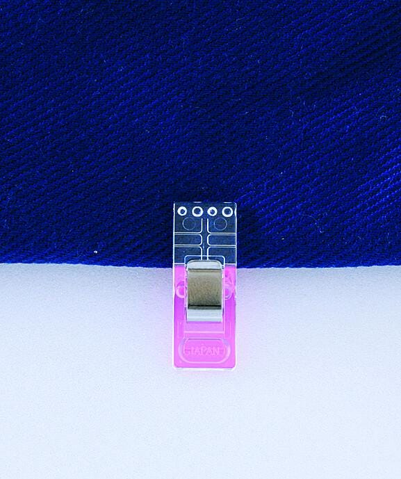 A clear plastic clip with a pink handle is attached to a blue fabric, with a white background.