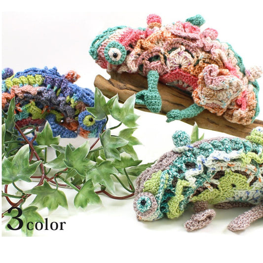 A crocheted chameleon toy in various colors, including pink, green, blue, and orange, is displayed alongside some green leaves.