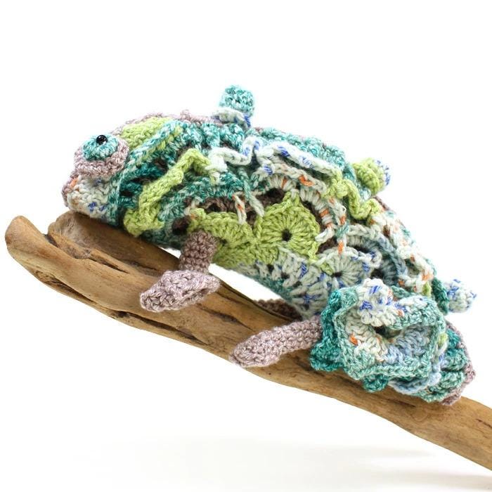 A crocheted chameleon figurine is perched on a wooden stick.