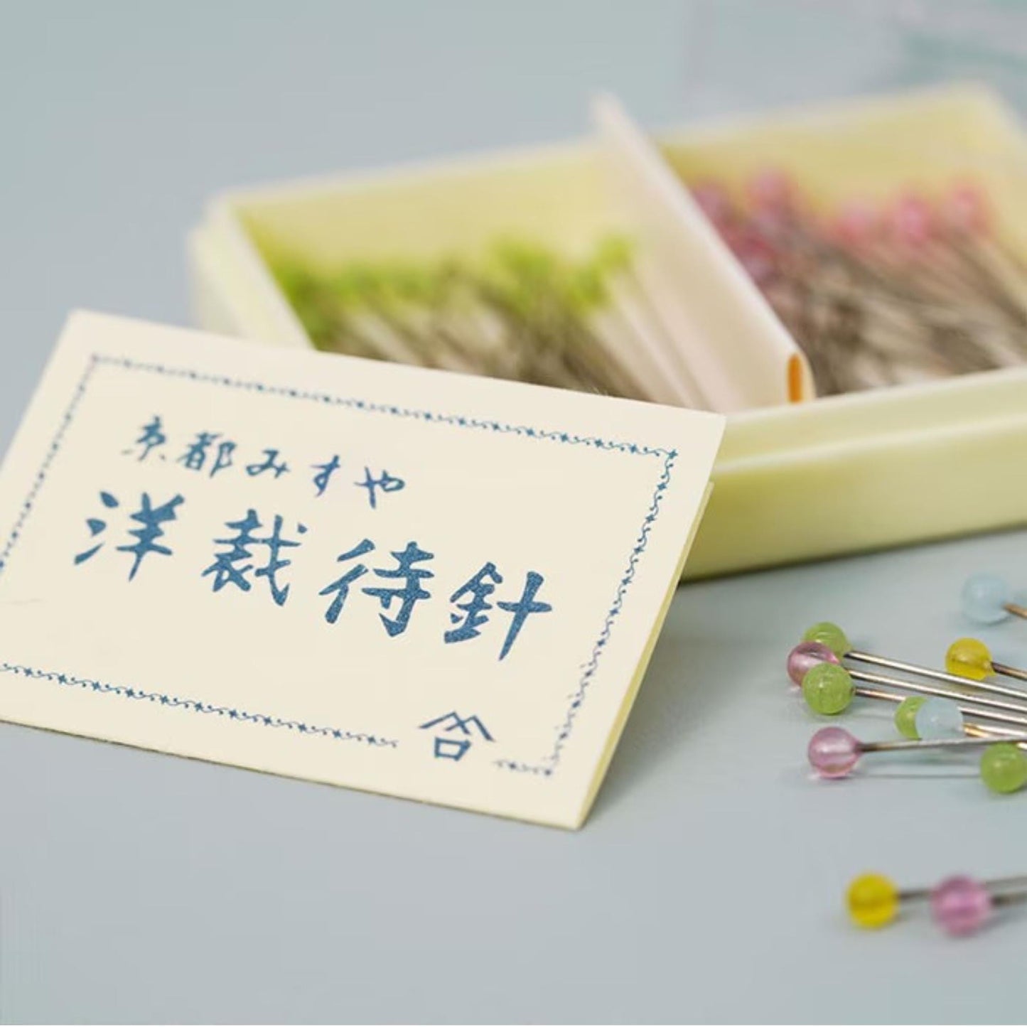 a close-up of a white card with blue text and a small symbol, alongside a box of colorful pins and needles on a light blue background.
