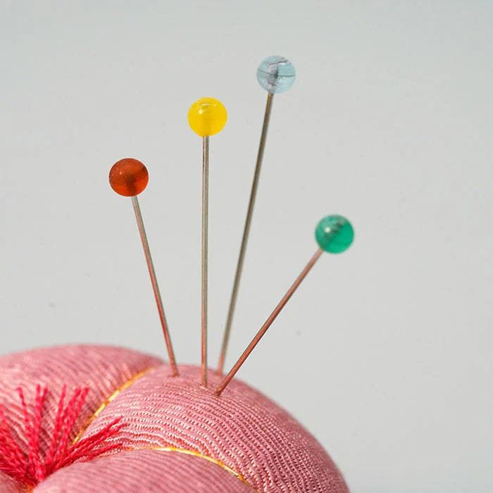 A close-up image of a pink fabric with colorful pins sticking out of it, against a plain white background.