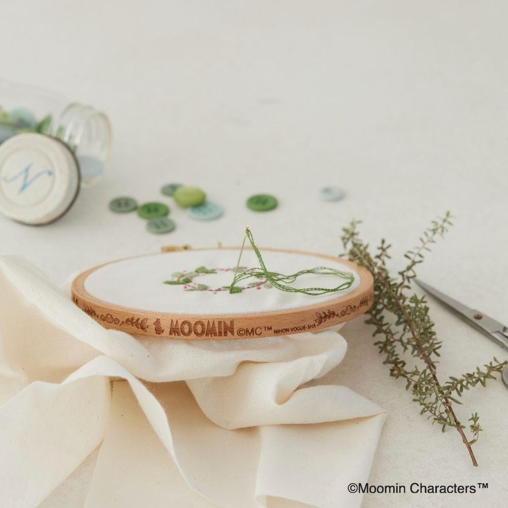 a wooden embroidery hoop with a piece of green thread on top, surrounded by various craft supplies and a sprig of greenery.