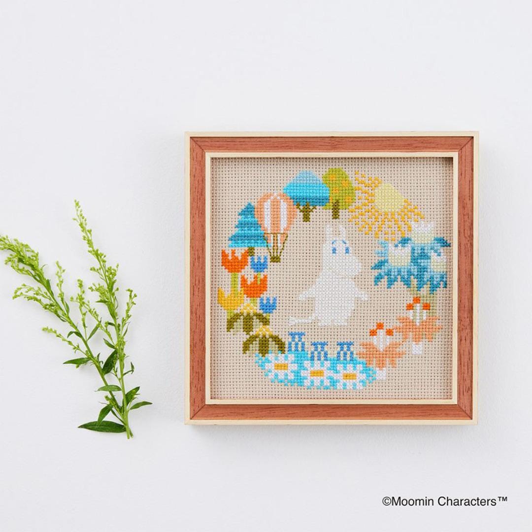 a cross-stitch design with various colorful elements, including a hot air balloon, flowers, and a small white character, all set against a white background.