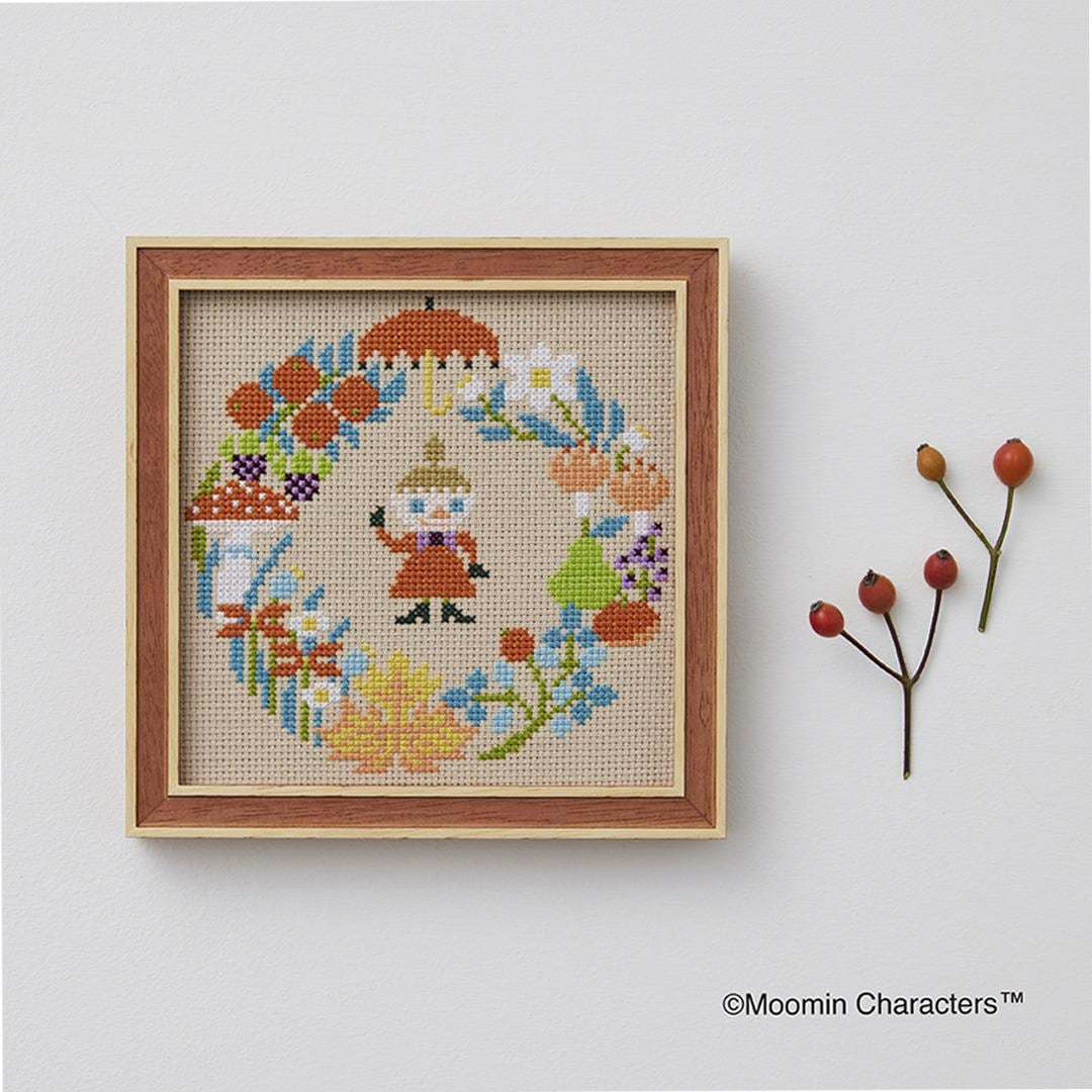 a cross-stitch design of a character, possibly a fairy or gnome, surrounded by various colorful flowers and leaves. The design is displayed in a wooden frame, and there are some small berries next to it.