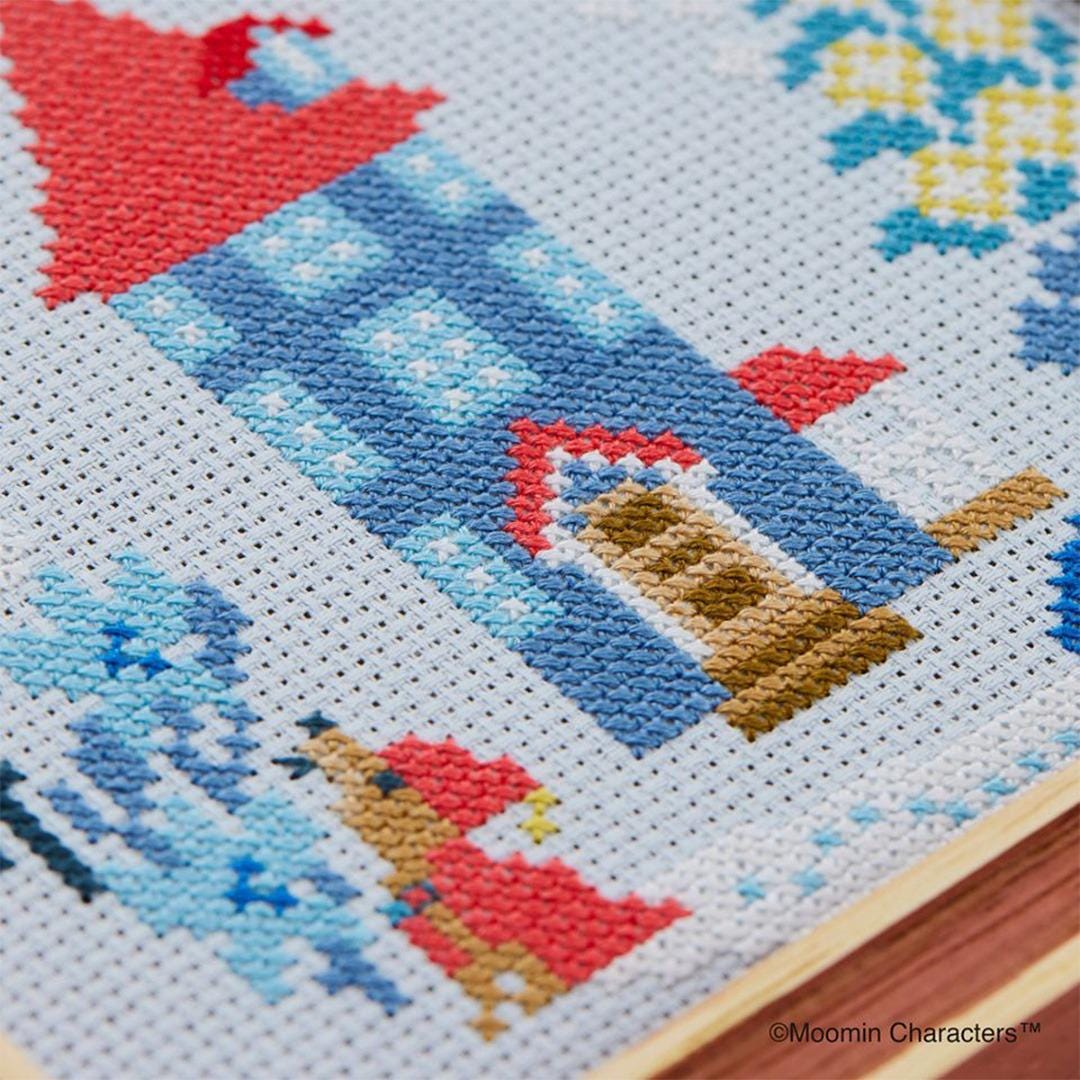 The image is a close-up of a cross-stitch pattern featuring a house, trees, and a dog. The pattern is detailed and colorful, showcasing the intricate work of cross-stitching.