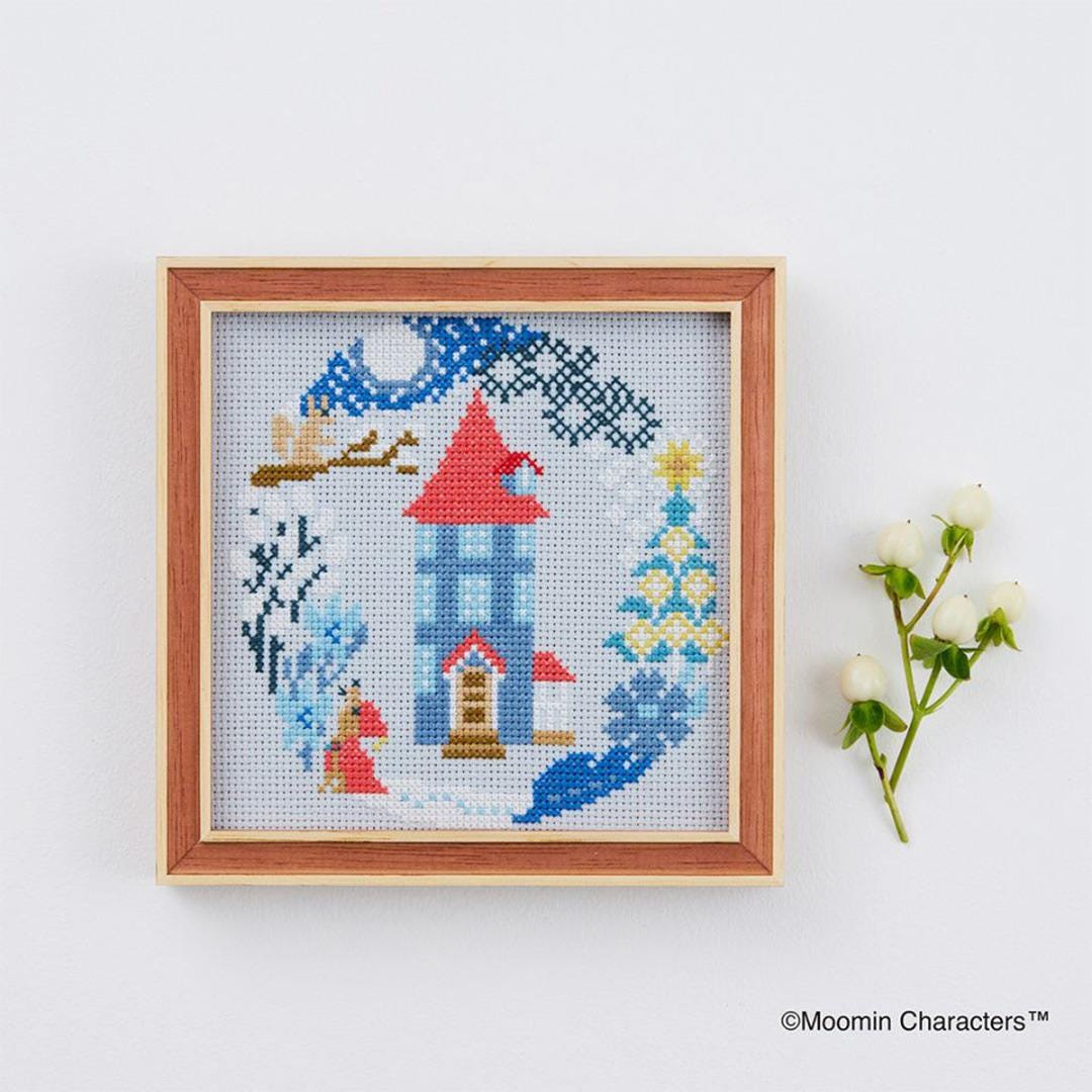 a cross-stitch design of a house, with a tree and other winter-themed elements. The design is framed and placed next to a branch of white flowers.